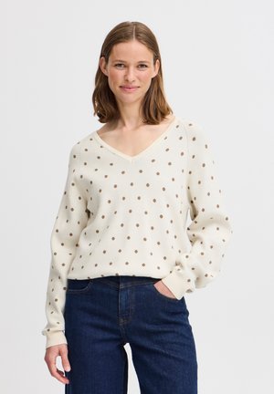 Pull crème à col en V avec des pois marron, présentant des manches longues et des poignets côtelés, associé avec un jean bleu foncé. Tissu tricoté texturé.