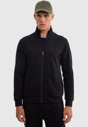 Sort zip-up sweatshirt lavet af blødt materiale, med høj krave, to sidelommer og ribbede manchetter. Båret med en grøn kasket.