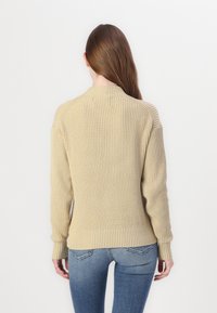 Suéter beige de punto con textura acanalada, cuello redondo y mangas largas. Combinado con jeans azules, mostrando una silueta ajustada.