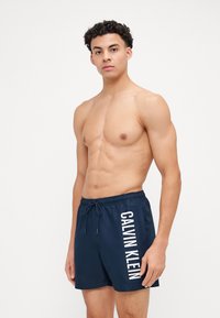 Marineblaue Badehosen mit einem verstellbaren Bund und einem weißen "CALVIN KLEIN" Logo an der Seite. Glattes, leichtes Material.
