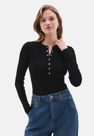 Black long-sleeve ribbed top with snap buttons along the neckline, paired with loose-fitting blue jeans. Smooth texture, fitted design.