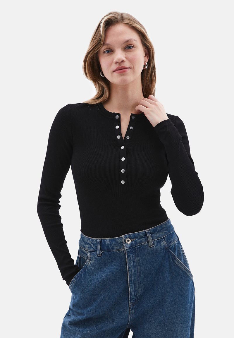 Schwarzes, langärmliges, geripptes Oberteil mit Druckknöpfen am Kragen, kombiniert mit locker sitzenden blauen Jeans. Glatte Textur, taillierter Schnitt.