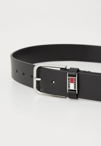 Zwarte leren riem met een zilveren metalen gesp. Heeft een rechthoekig logo met rode en witte accenten. Vier verstelbare gaten.