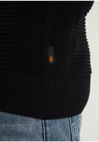 Pull noir en tricot avec une texture côtelée et une petite étiquette orange contrastante sur le côté. Associé à un jean en denim bleu clair.