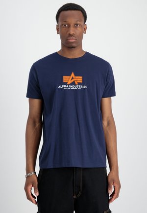 Tamno plavi pamučni majica kratkih rukava s narančastim grafičkim logom i natpisom "ALPHA INDUSTRIES" na prsima.