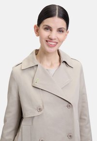 Beige trenchcoat met een dubbelzijdig ontwerp, grote kraag en een extra groene accent, gemaakt van een gladde, duurzame stof.