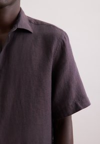 Korte mouwen, geknoopte shirt van donkerpaarse linnen. Kenmerkt zich door een ontspannen pasvorm met een subtiele textuur en nette stiksels.