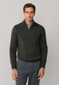 Donkergrijze zip-up trui met een hoge kraag, gemaakt van zacht materiaal. Gelaagd over een blauw-wit gestreept shirt, gedragen met grijze broek.