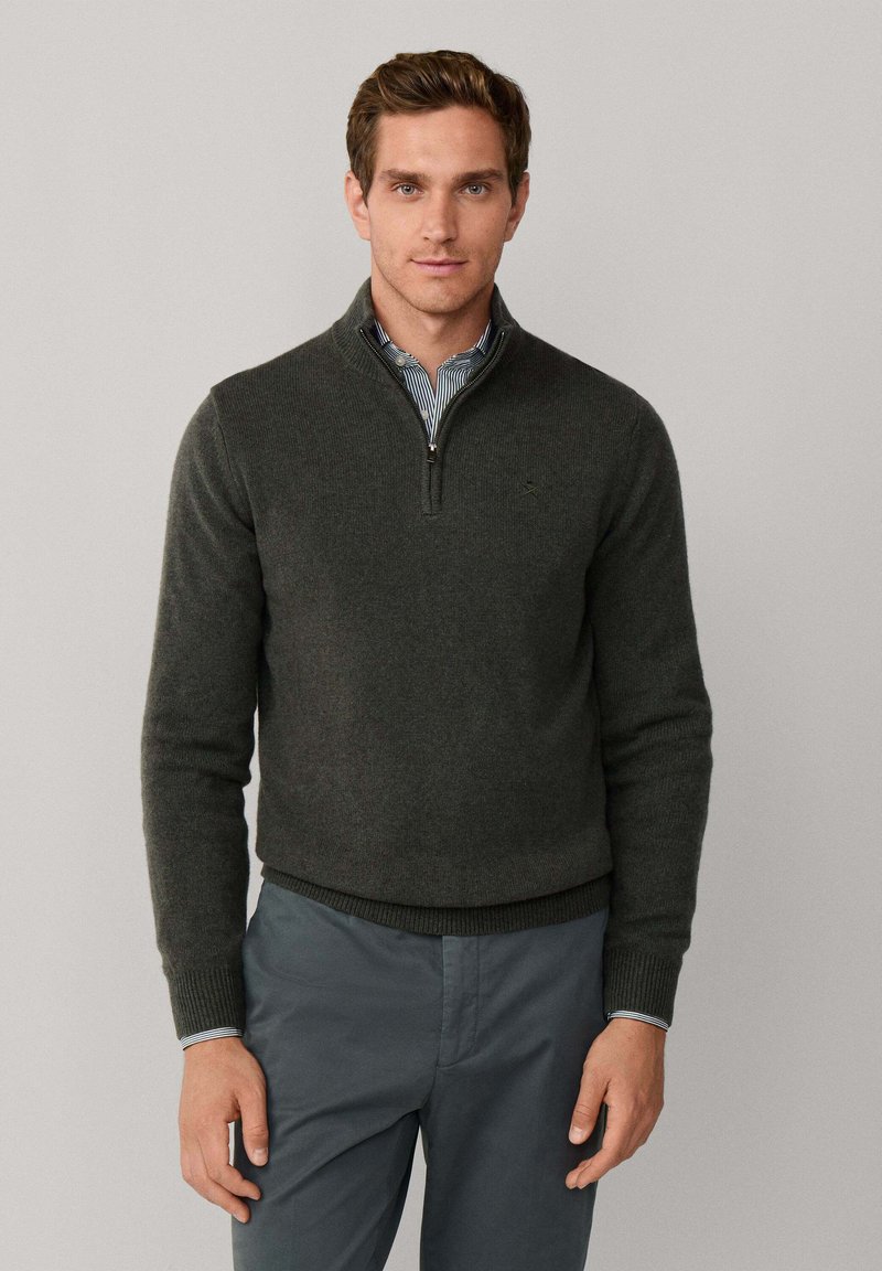 Donkergrijze zip-up trui met een hoge kraag, gemaakt van zacht materiaal. Gelaagd over een blauw-wit gestreept shirt, gedragen met grijze broek.