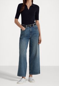 Polo shirt navy, jeans a vita alta a gamba larga in blu con lavaggio chiaro e scarpe da ginnastica bianche. I jeans presentano grandi tasche e una vestibilità comoda.
