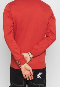 Sweat-shirt à capuche rouge avec manches longues, col rond et poignets côtelés. Présente une texture lisse et un design simple de couleur unie.