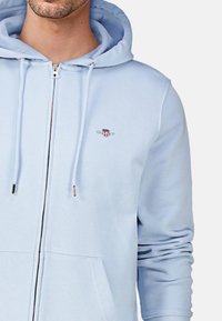 Svetlomodrá mikina na zips vyrobená z mäkkého materiálu. Obsahuje predné vrecko, nastaviteľné šnúrky a vyšívané logo na hrudi.