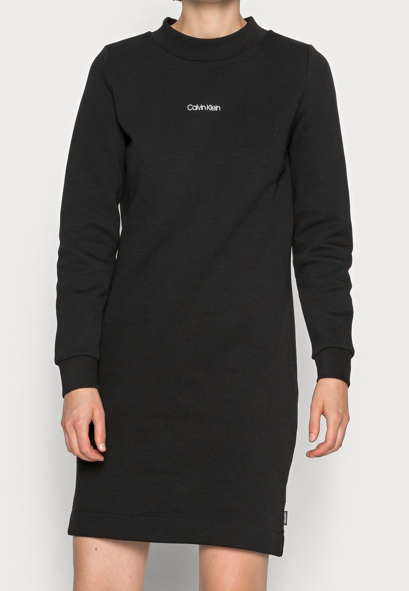 Robe décontractée noire à manches longues avec col rond, poignets ajustés et petit logo blanc "Calvin Klein" sur la poitrine, portée par une personne debout.