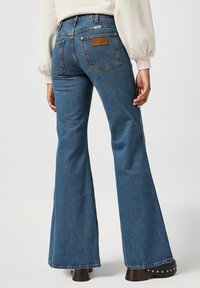 Light blue flared jeans featuring a high waist, rear pockets with a leather logo patch, and a subtle faded texture. Worn with brown heeled boots.