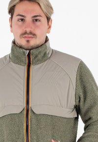 Giacca in pile con la parte superiore tan e il corpo verde oliva, caratterizzata da un collo alto, tessuto strutturato, zip frontale e un discreto logo sul petto.