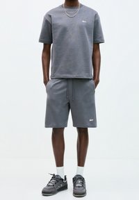 T-shirt gris en coton mélangé à manches courtes avec un col rond, associé à un short gris assorti. Les deux pièces présentent des logos brodés. Porté avec des baskets noires.