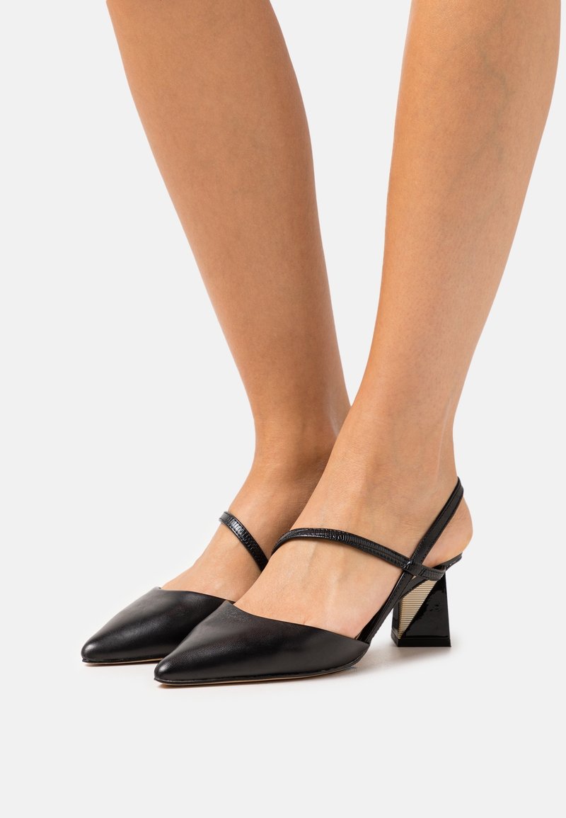Jambes nues portant des escarpins slingback noirs à bout pointu avec fines brides chevilles et talons carrés sur un fond clair uni.
