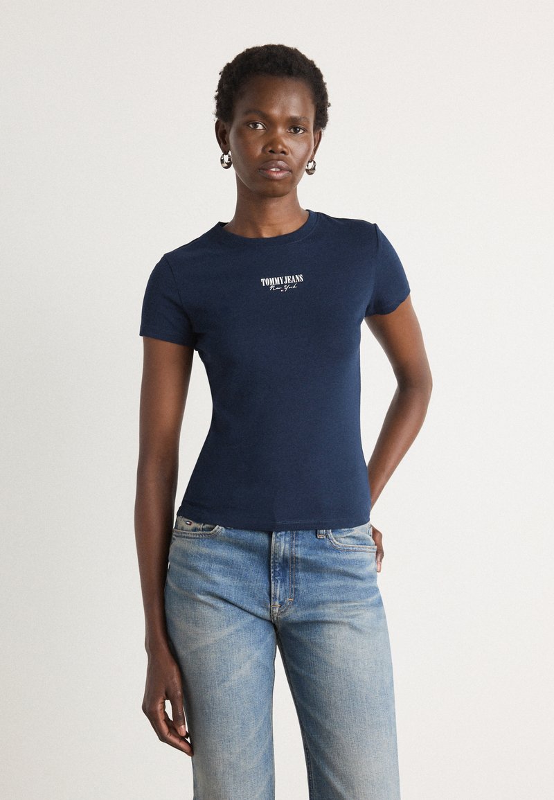Tamsiai mėlynas medvilninis marškinėliai su prigludusia forma ir trumpomis rankovėmis. Krūtinėje yra baltas "TOMMY JEANS" logotipas. Sujungti su šviesiai mėlynomis džinsais.