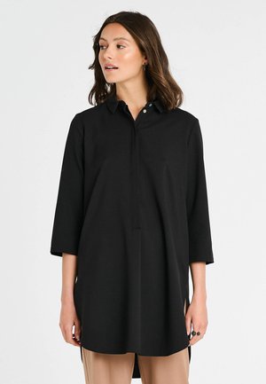 Tunique noire avec col, manches trois-quarts, détails de boutons sur le devant et fentes sur les côtés. Fabriquée en tissu lisse.