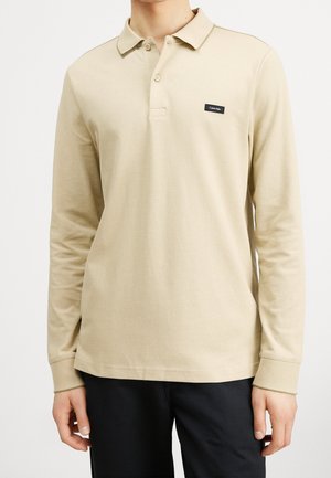 Polo beige à manches longues avec col et deux boutons, patch logo Calvin Klein noir sur la poitrine, porté avec un pantalon noir.