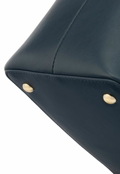 Navy leather handbag corner featuring brass hardware, smooth texture, and contrast stitching, with rounded edges and visible seam detail.