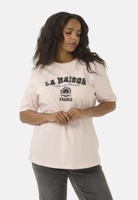 T-shirt en coton rose clair à manches courtes avec un texte graphique noir "LA MAISON" et "FRANCE" à l'avant. Design simple et décontracté.