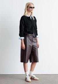 Schwarzer gestrickter Cardigan über einem hellblauen Hemd mit Kragen, kombiniert mit braunen Leder-Shorts und beigen Sneakers mit weißen Socken.