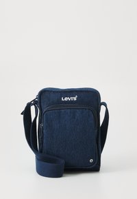 Plavi traper torbica preko ramena s patentnim zatvaračem na prednjoj strani, podesivim remenom i Levi's logom na vrhu. Glatka tekstura i pravokutni oblik.