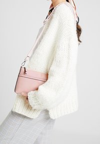 Valkoinen neulepusero, jossa on reilut hihat, yhdistettynä pieneen vaaleanpunaiseen käsilaukkuun, jossa on olkahihna ja vetoketjukiinnitys.