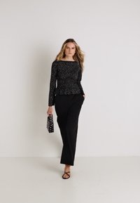Crna majica dugih rukava s peplum dizajnom i mrljastim uzorkom, uparena s labavim crnim hlačama. Model drži malu torbicu s točkicama.