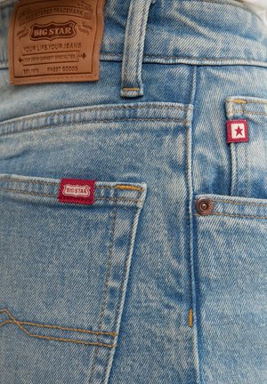 Gai zila džinsu bikses ar brūnas ādas zīmola uzlīmi, sarkaniem akcentu tagiem un priekšējo kabatu ar redzamām dūriena detaļām.