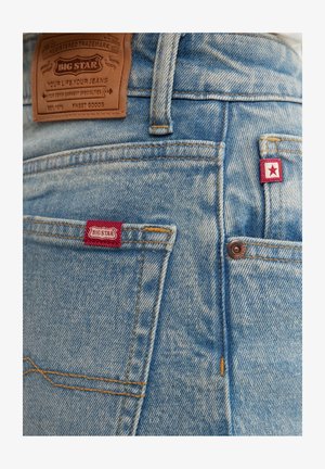 Heled sinised teksad pruunist nahast brändi sildiga, punaste aktsentetikettidega ning esiküljel nähtavad õmblusdetailid.