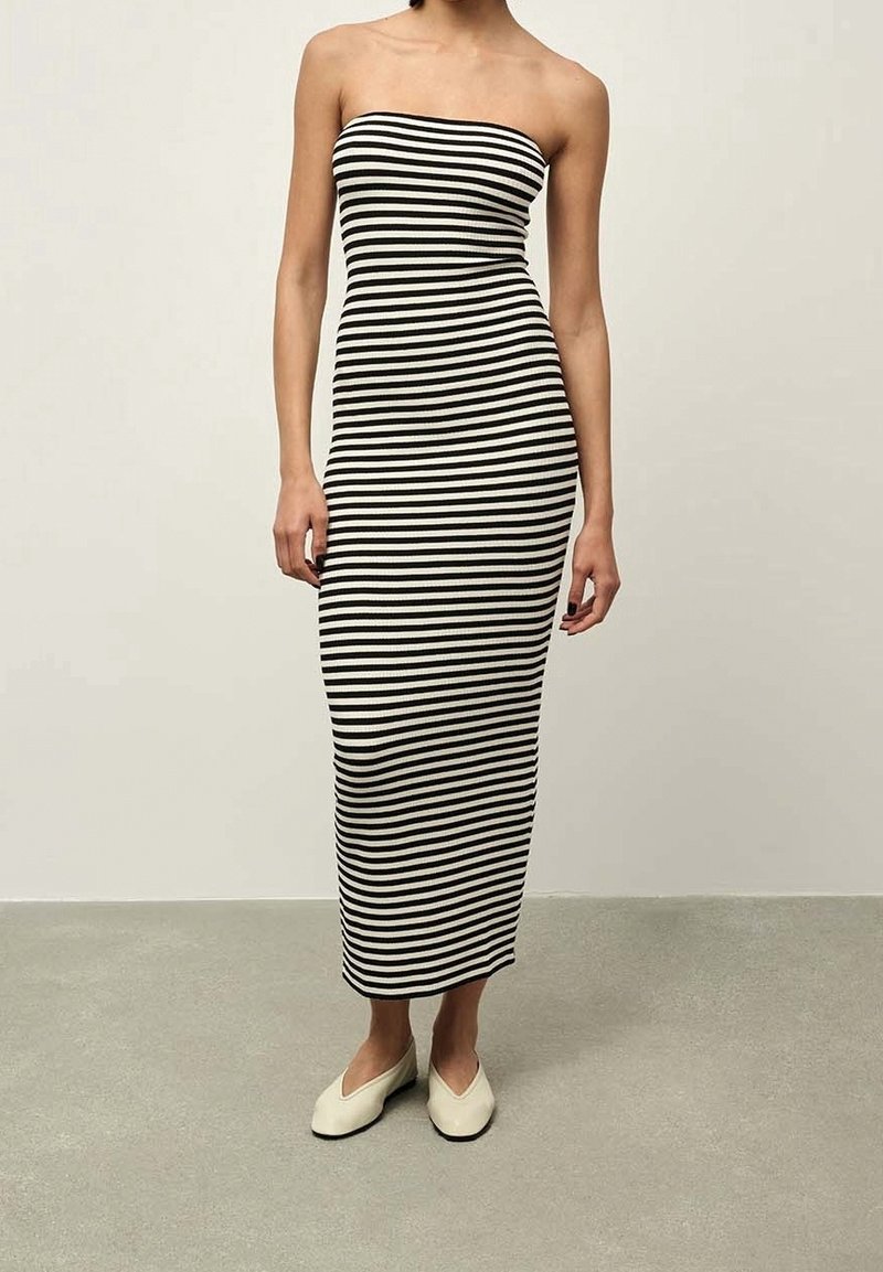 Femme portant une robe bustier rayée horizontalement noire et blanche et des ballerines blanches à bouts pointus, debout sur un sol gris devant un fond beige.