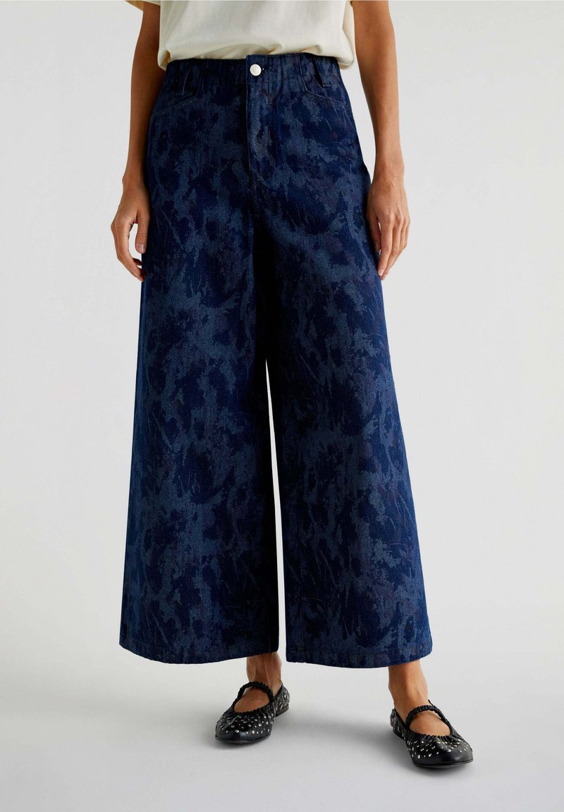 Pantalons larges en denim foncé avec ceinture élastique, grandes poches et un motif texturé subtil. Associés à des ballerines noires.