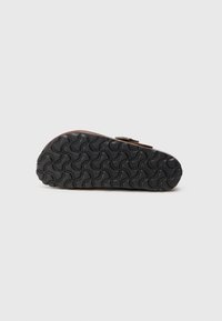 Sandal maro cu o talpă din cauciuc negru texturat, având un model în formă de val și un detaliu cu cataramă central. Partea de jos este prezentată în mod proeminent.