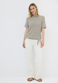 Gestreiftes, kurzärmeliges Oberteil in Creme und Schwarz, mit einer Bindeschleife an der Seite, kombiniert mit weißen, weit geschnittenen Hosen und braunen Sandalen.