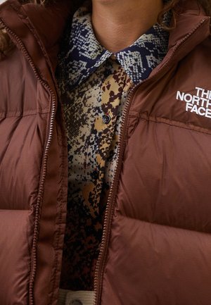 Bruine pufferjas met het "The North Face" logo, gedragen over een veelkleurig, patroonrijk overhemd met een blauw-witte kraag.