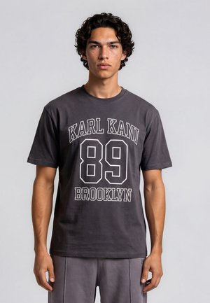Junger Mann mit lockigem dunklem Haar trägt ein dunkelgraues T-Shirt mit weißem Aufdruck "KARL KANI 89 BROOKLYN" und steht vor einem einfarbigen Hintergrund.