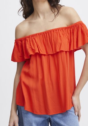 Vrouw draagt een feloranje off-shoulder blouse met ruches, gecombineerd met lichtblauwe spijkerbroek, staand tegen een effen achtergrond.