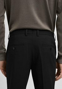Schwarze maßgeschneiderte Hosen mit glatter Oberfläche, Gürtelschlaufen und zwei Knopf-Gesäßtaschen. Getragen mit einem hellbraunen Pullover.