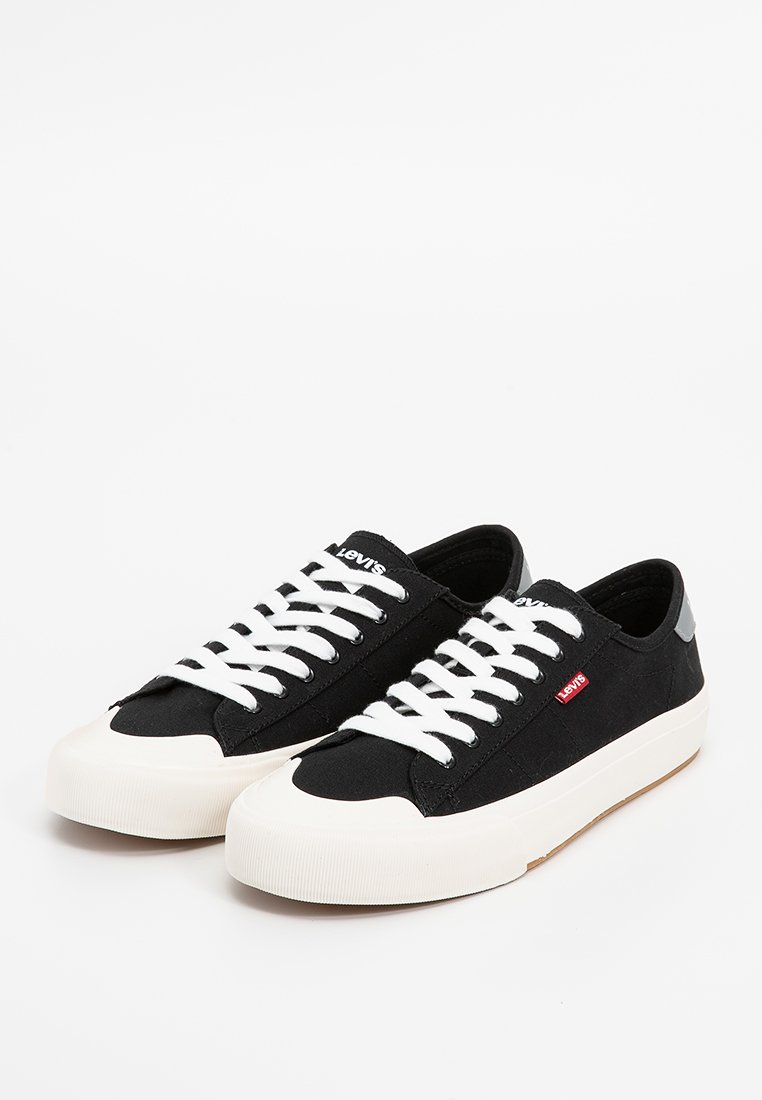 Zapatillas de lona negras con cordones blancos y suela de goma. Presentan una etiqueta de logo roja y un detalle gris en la parte trasera. Diseño de punta redonda.