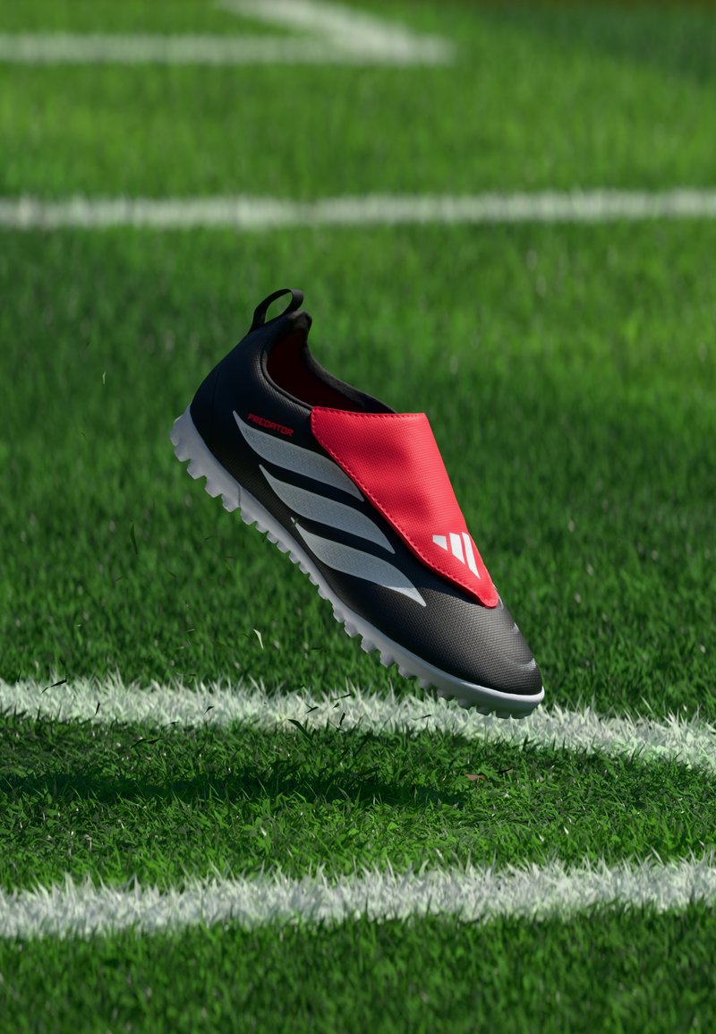 Chaussure de football Adidas Predator noire et rouge flottant au-dessus de lignes blanches sur un terrain en gazon artificiel vert.