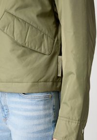 Giacca verde oliva con finitura texturizzata, dotata di tasca laterale e etichetta del marchio visibile sull'orlo, abbinata a jeans azzurri.