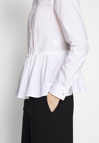 Donna che indossa una blusa peplum bianca con maniche lunghe e polsini con bottoni, con una mano nella tasca dei pantaloni neri, mostrata dalla vita a metà coscia.