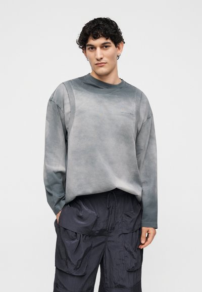 Νέος άνδρας με κατσαρά μαλλιά στέκεται με τα χέρια στις τσέπες ενός σκούρου cargo παντελονιού, φορώντας γκρι μακρυμάνικη μπλούζα σε απλό φόντο.