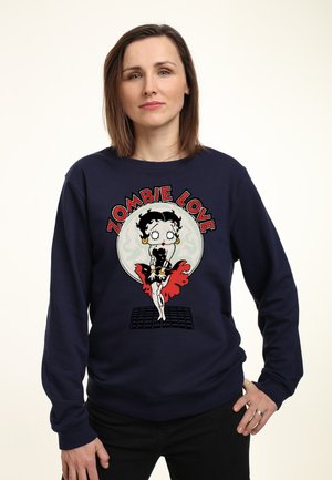 Kvinde med skulderlangt brunt hår iført en marineblå sweatshirt med en zombie tegneseriefigur og tekst "Zombie Love" mod en lys baggrund.