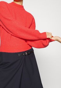 Pull en maille côtelée rouge avec col montant, manches longues. Associé à une jupe foncée avec une ceinture noire et des détails en métal argenté.