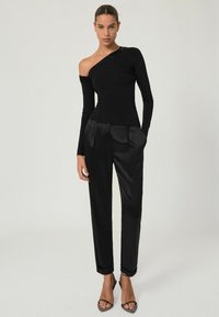 Žena oblečená v čiernom top s jedným ramenom a dlhým rukávom, ozdobeným flitrami, a v čiernych saténových nohaviciach s vysokým pásom, stojí so zahnutými rukami v vreckách.