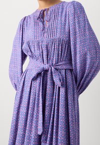 Lilla kjole med pink mønster, lange pufærmer, rynket krop, læg, bindebånd i taljen og flydende skørt. Tekstureret stof med blød fald.