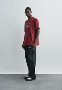 Κόκκινο γραφικό t-shirt με εκτύπωση "ACDS", μαύρα χαλαρά παντελόνια με πλαϊνές φερμουάρ, μαύρα παπούτσια με λευκές λεπτομέρειες και γυαλιά ηλίου.